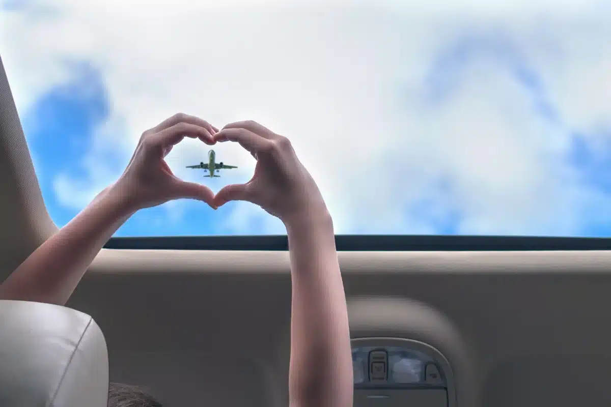Person making a heart shape with their hands around a passing airplane in a blue sky, viewed from inside a car.