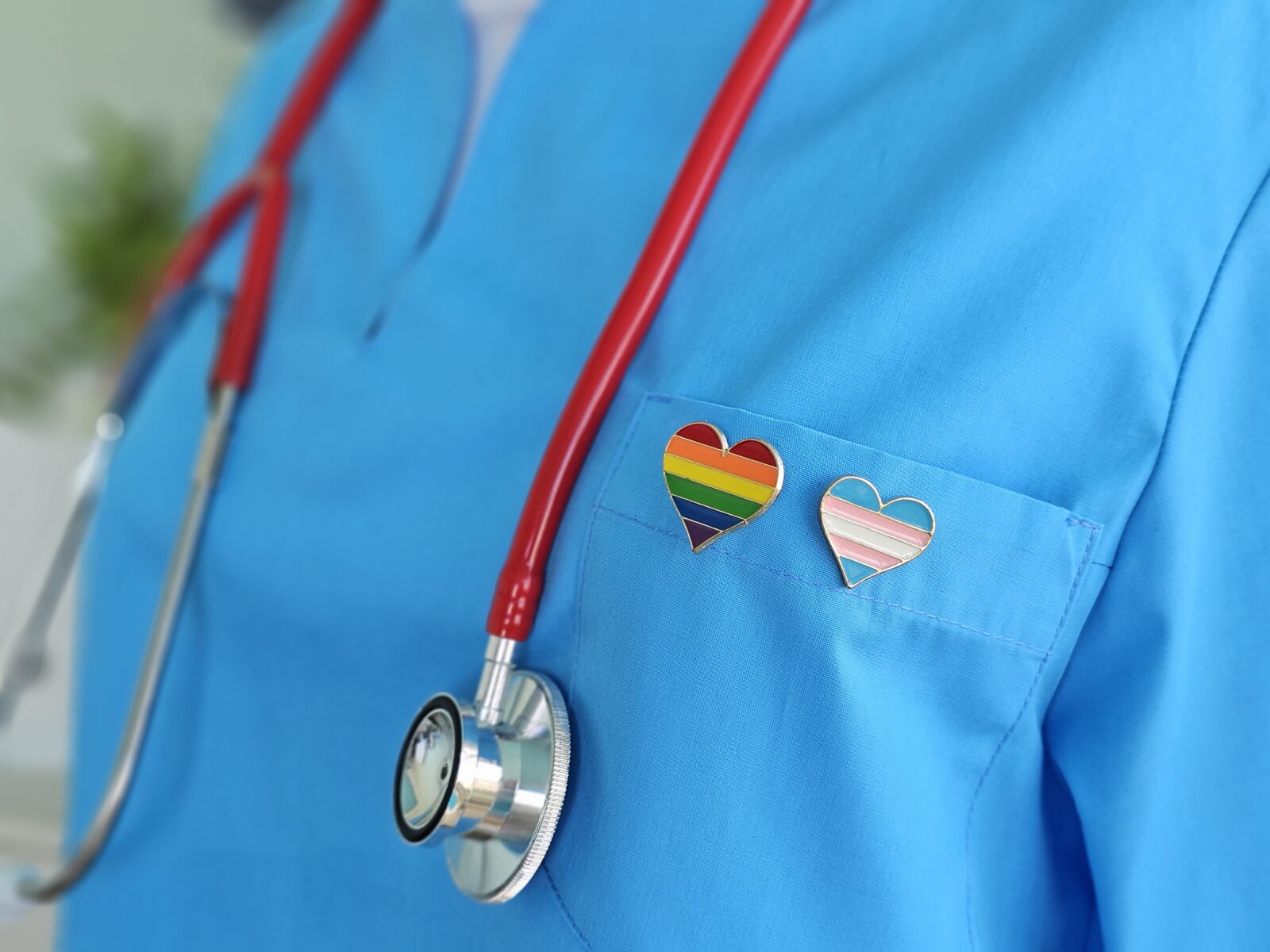 Healthcare worker wearing blue scrubs with a rainbow heart pin and a red stethoscope.