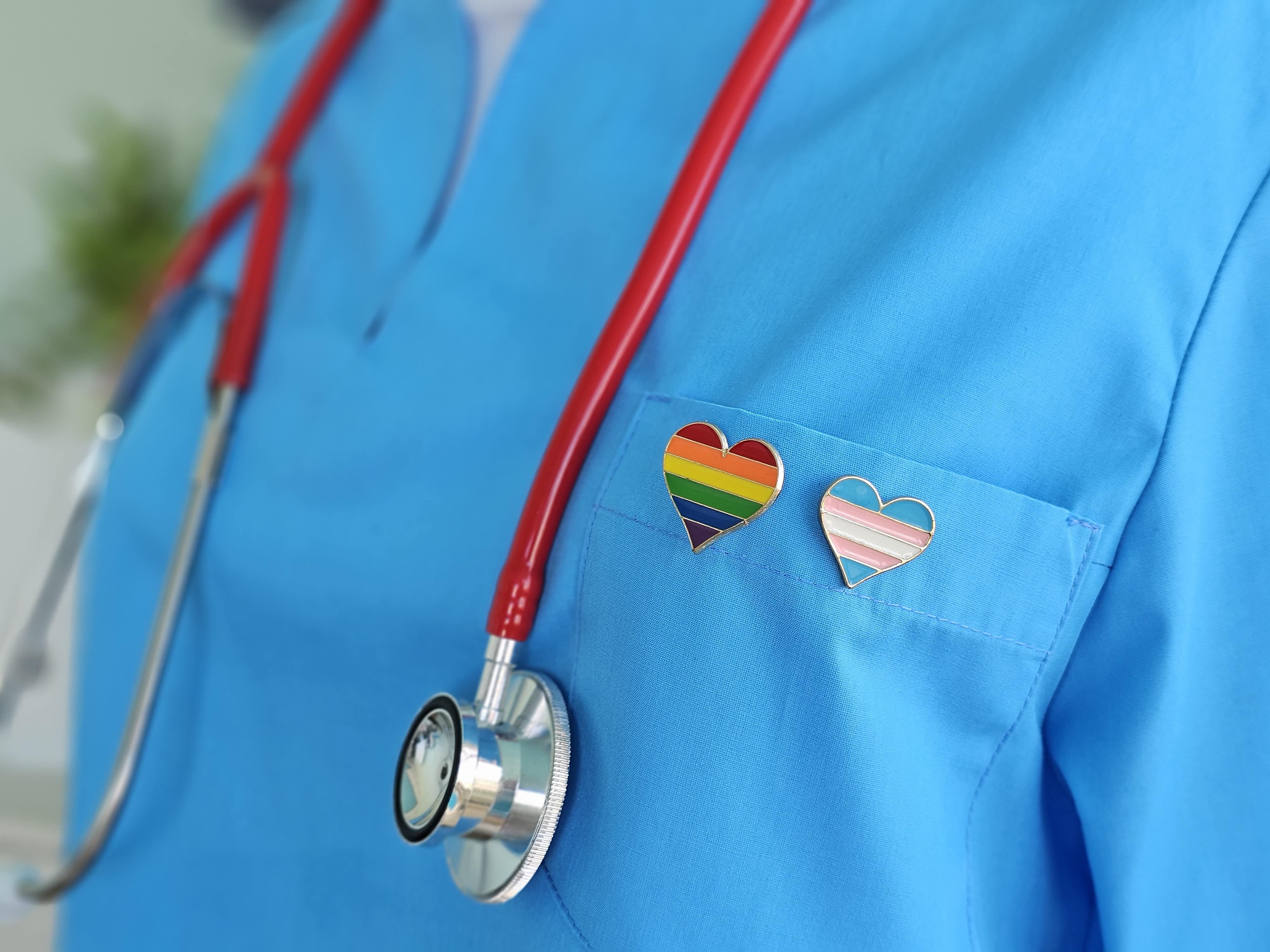 Un professionnel de santé portant une blouse bleue, ornée d'un pin's en forme de cœur arc-en-ciel et d'un stéthoscope rouge.